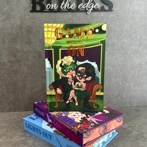 Peut inclure: Des livres empilés avec des couvertures colorées. Le livre du dessus est intitulé "GAME ON" avec un design vert et or et un couple de dessins animés. En dessous, les livres sont intitulés "LIGHTS OUT" et "CAL". Le nom de l'auteur, "NAVESSA ALLEN", est sur les dos.