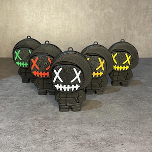 Könnte beinhalten: Sechs schwarze Figuren mit Kapuzen und leuchtenden Gesichtern mit genähten Mündern. Jede Figur hat eine andere Farbe für Augen und Mund: grün, rot, weiß, gelb. Die Figuren stehen auf einer grauen Oberfläche.