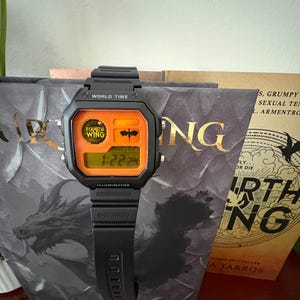 May include: A black digital watch with a rectangular face and an orange display panel. The watch features the text "WORLD TIME" and "ILLUMINATOR" on the bezel, with "FOURTH WING" and a dragon silhouette graphic printed on the orange screen area.