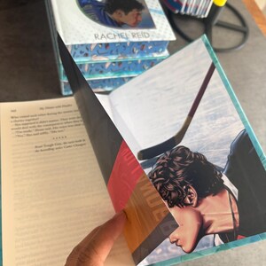 May include: A stack of books titled "Rivalry" by Rachel Reid. The open book reveals an illustration of a hockey player. The cover features two male figures in hockey uniforms. The book's pages are edged in teal.