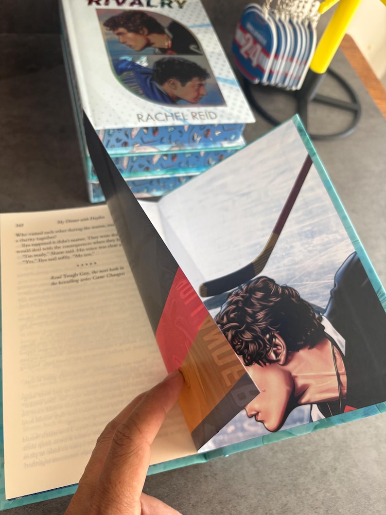 May include: A stack of books titled "Rivalry" by Rachel Reid. The open book reveals an illustration of a hockey player. The cover features two male figures in hockey uniforms. The book's pages are edged in teal.