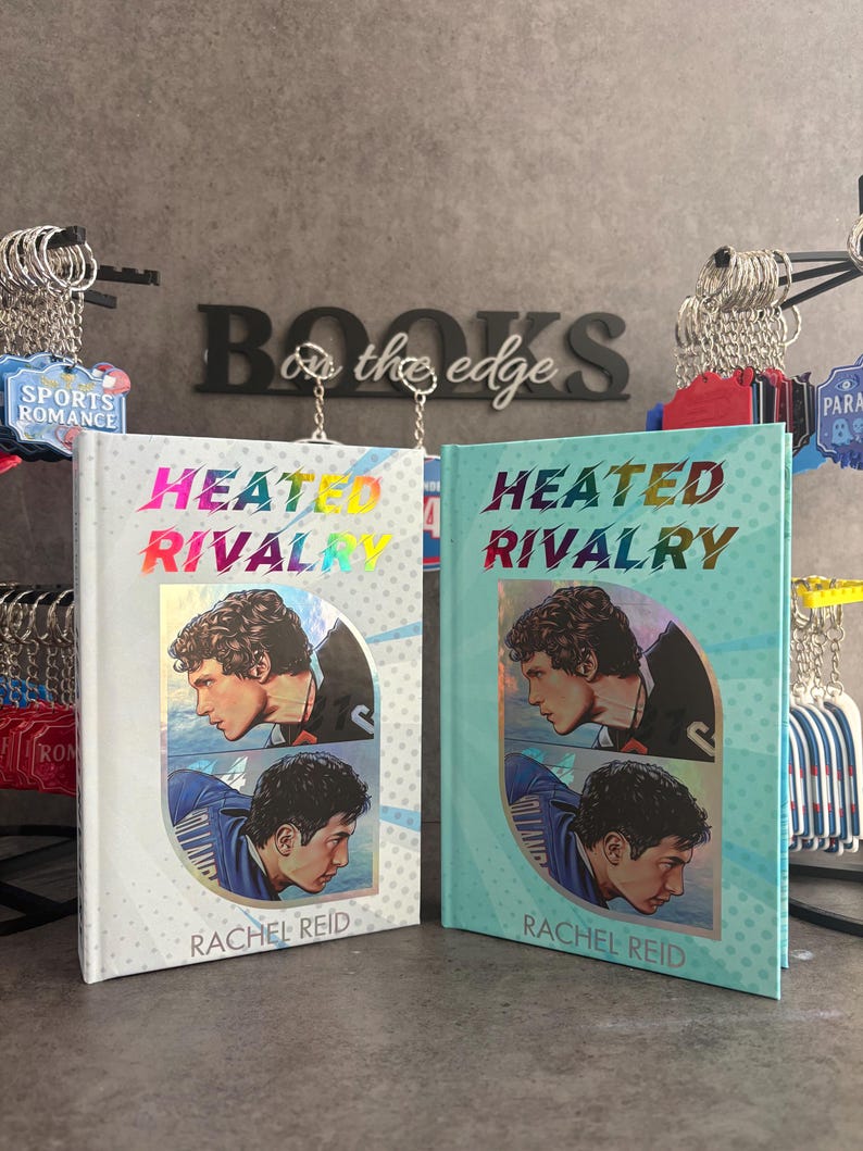 May include: Two hardcover books titled "Heated Rivalry" by Rachel Reid are displayed. One book has a white cover with a holographic title, and the other has a teal cover. Both feature a portrait of a man. The background includes keychains and the words "Books on the edge."