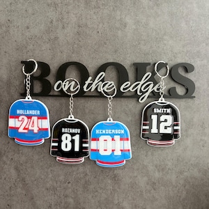 Peut inclure: Quatre porte-clés en forme de maillots de hockey sont suspendus à une enseigne noire avec l'inscription "BOOKS on the edge". Les porte-clés représentent des maillots de hockey avec des noms et des numéros. Les maillots sont bleus, noirs et blancs.