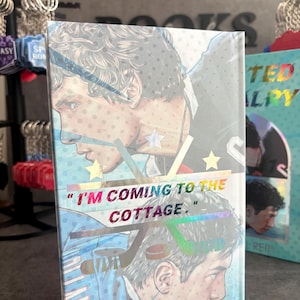 May include: A book with a holographic cover featuring a male illustration in a hockey uniform. The text "I'M COMING TO THE COTTAGE" is displayed in a colorful, iridescent font. The book has a light blue background.