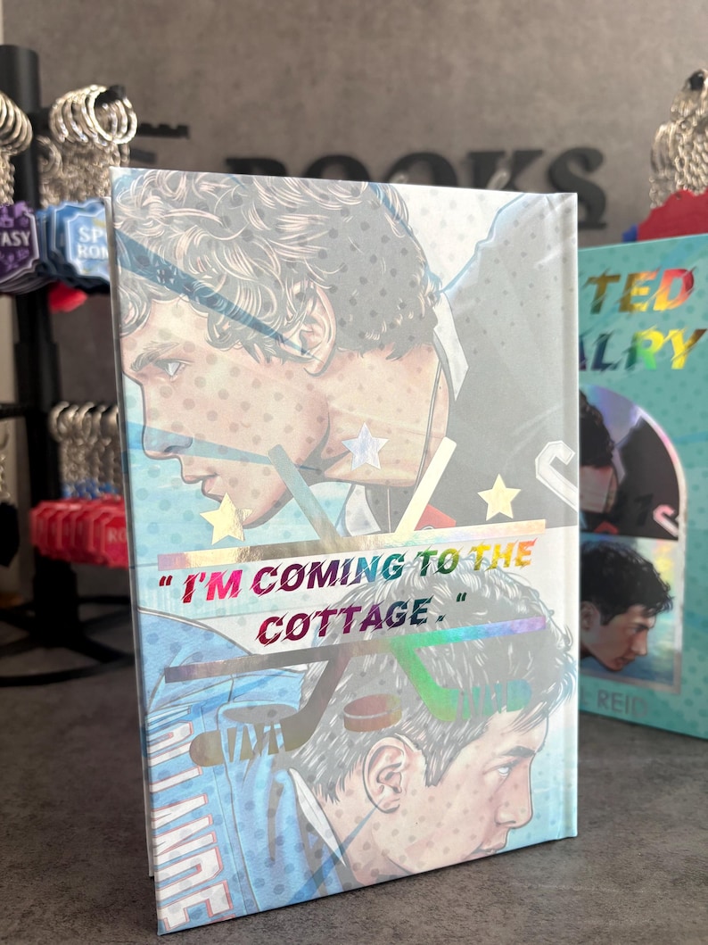 May include: A book with a holographic cover featuring a male illustration in a hockey uniform. The text "I'M COMING TO THE COTTAGE" is displayed in a colorful, iridescent font. The book has a light blue background.