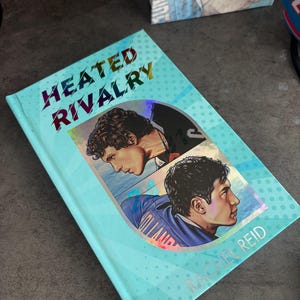May include: A hardcover book titled "Heated Rivalry" by Rachel Reid. The cover is light blue with a pattern of small white dots. It features two male portraits and the title in holographic lettering.