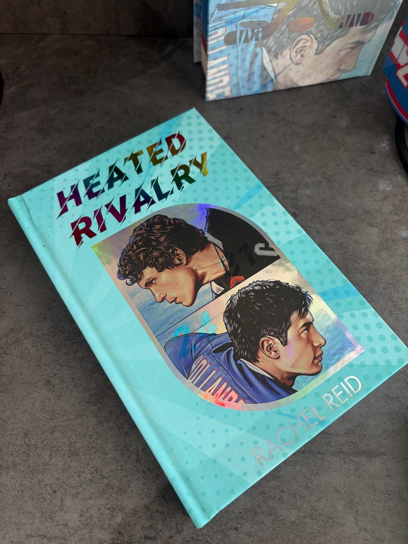 May include: A hardcover book titled "Heated Rivalry" by Rachel Reid. The cover is light blue with a pattern of small white dots. It features two male portraits and the title in holographic lettering.