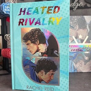 May include: A hardcover book titled "Heated Rivalry" by Rachel Reid. The cover is light blue with a rainbow-colored title and illustrations of two men. The book has a light blue background with small white polka dots.