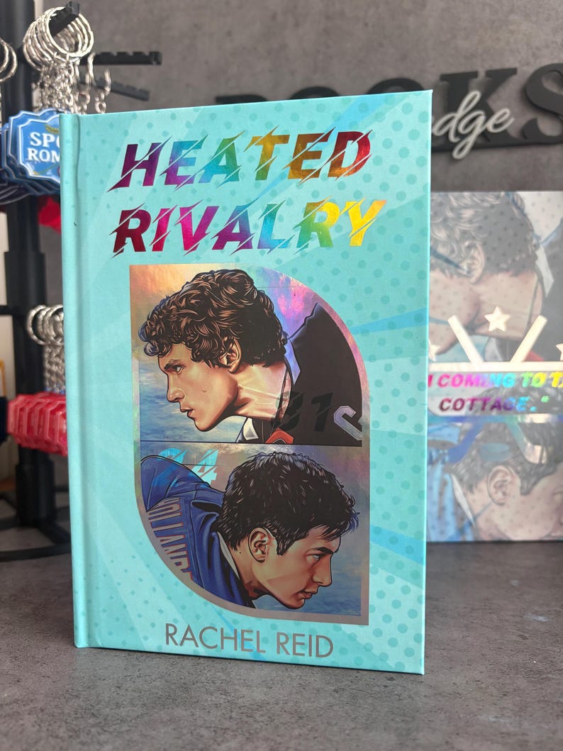 May include: A hardcover book titled "Heated Rivalry" by Rachel Reid. The cover is light blue with a rainbow-colored title and illustrations of two men. The book has a light blue background with small white polka dots.