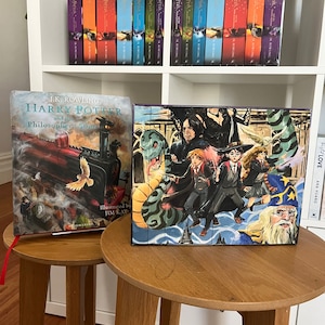 May include: A collection of Harry Potter books, including a boxed set and individual volumes, displayed on a white shelf and two wooden side tables. The books feature colorful covers with illustrations and the title "Harry Potter".