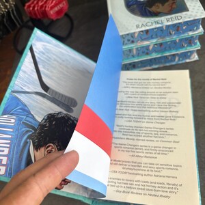May include: A stack of books titled "Heated Rivalry" by Rachel Reid. The cover features two people in hockey jerseys. The open book reveals a hockey player illustration and text. The book's pages have a blue and white patterned edge.