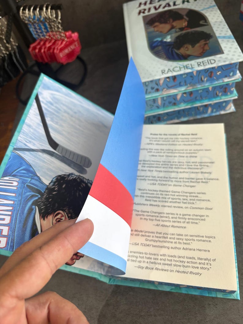 May include: A stack of books titled "Heated Rivalry" by Rachel Reid. The cover features two people in hockey jerseys. The open book reveals a hockey player illustration and text. The book's pages have a blue and white patterned edge.