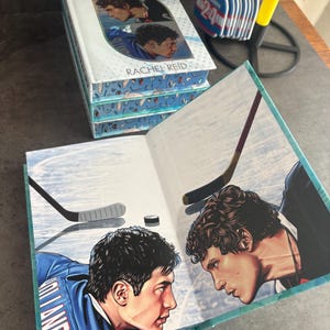 May include: A stack of books titled "Heated Rivalry" by Rachel Reid. The book cover features two men in hockey uniforms. The open book shows a hockey scene with two players facing each other, hockey sticks, and a puck.