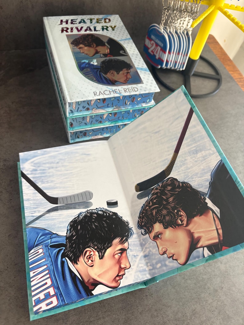 May include: A stack of books titled "Heated Rivalry" by Rachel Reid. The book cover features two men in hockey uniforms. The open book shows a hockey scene with two players facing each other, hockey sticks, and a puck.