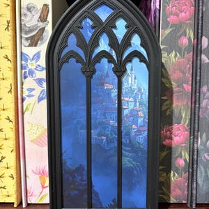 Könnte beinhalten: Ein schwarzer Buchstütze im gotischen Stil mit einem detaillierten Fensterdesign. Das Fenster zeigt eine malerische Landschaft mit einem Schloss und einer Stadt auf einer Klippe. Die Buchstütze steht zwischen Büchern mit floralen und naturnahen Einbänden.