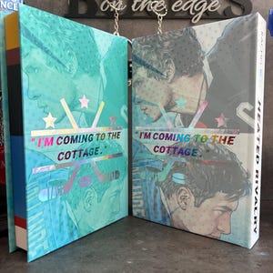 May include: Two hardcover books with teal and blue covers featuring a male portrait and the text "I'M COMING TO THE COTTAGE." The book on the right has the title "HEATED RIVALRY" on the spine. The background includes the word "BOOKS" and "on the edge."