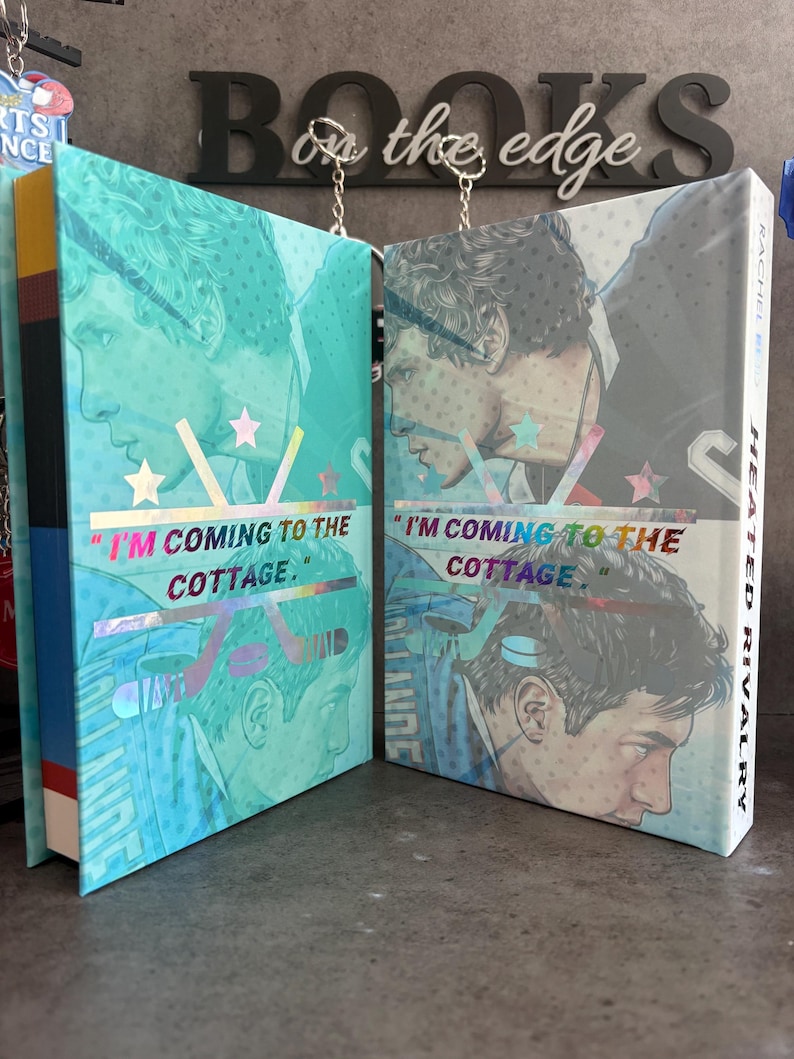 May include: Two hardcover books with teal and blue covers featuring a male portrait and the text "I'M COMING TO THE COTTAGE." The book on the right has the title "HEATED RIVALRY" on the spine. The background includes the word "BOOKS" and "on the edge."