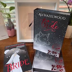 Może przedstawiać: Ułożone w stos książki w twardej oprawie zatytułowane "Bride" autorstwa Ali Hazelwood. Książki mają czarną okładkę z czerwonymi i białymi literami oraz ilustracjami wilka i kobiety. Leżą na drewnianej powierzchni, z oprawionym rysunkiem i kwiatami w tle.