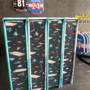 May include: Four decorative bookends with a black background and a pattern of hockey-related graphics in white, red, and blue. The edges are trimmed with a teal and white pattern. Two hockey jersey keychains hang above the bookends.