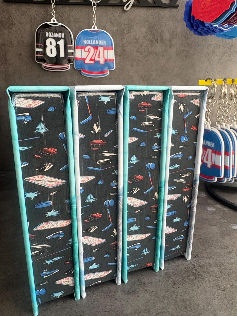May include: Four decorative bookends with a black background and a pattern of hockey-related graphics in white, red, and blue. The edges are trimmed with a teal and white pattern. Two hockey jersey keychains hang above the bookends.