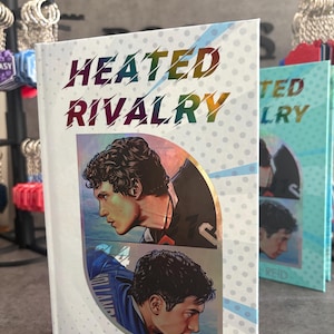 May include: A book titled "Heated Rivalry" by Rachel Reid. The cover features two male portraits in profile, with the title in rainbow gradient lettering. The background is white with a dotted pattern. Other books are visible in the background.