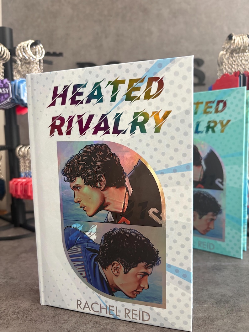 May include: A book titled "Heated Rivalry" by Rachel Reid. The cover features two male portraits in profile, with the title in rainbow gradient lettering. The background is white with a dotted pattern. Other books are visible in the background.