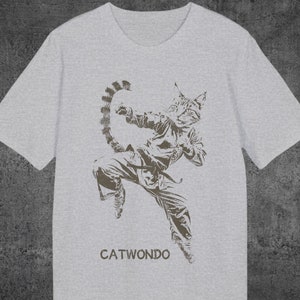 Puede incluir: Una camiseta gris con un gráfico de un gato haciendo una pose de artes marciales. El gato lleva un uniforme de karate y tiene una cola que se enrolla detrás de él. El texto "CATWOND0" está impreso debajo del gráfico.