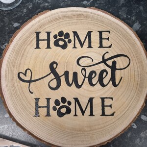 以下が含まれることがあります： 黒い文字で「Home Sweet Home」と書かれた丸い木製の看板。 「Home」という単語は、両側に肉球の跡で飾られています。
