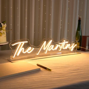 Könnte beinhalten: Ein individuelles Neonschild mit dem Schriftzug "The Martins" in einer warm leuchtenden, gelben Schreibschrift. Das Schild ist auf einer transparenten, konturierten Acrylplatte montiert und steht auf einer ebenen Fläche, die sanft beleuchtet wird.