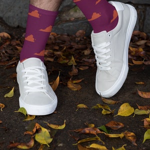 May include: A pair of white sneakers with purple socks featuring an orange outline of the state of Virginia. The socks are worn by a person standing on a surface covered in fallen leaves.