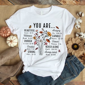 May include: White t-shirt with a floral design and the text "YOU ARE..." with inspirational quotes and bible verses. The text includes "Beautiful", "Victorious", "Enough", "Created", "Strong", "Amazing", "Capable", "Chosen", "Never Alone", and "Always Loved".