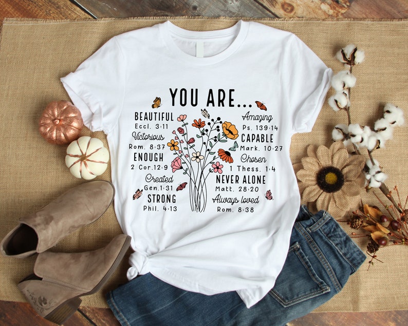 May include: White t-shirt with a floral design and the text "YOU ARE..." with inspirational quotes and bible verses. The text includes "Beautiful", "Victorious", "Enough", "Created", "Strong", "Amazing", "Capable", "Chosen", "Never Alone", and "Always Loved".