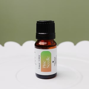 Puede incluir: Una pequeña botella de vidrio ámbar con una tapa negra, que contiene aceite esencial. La etiqueta es blanca con acentos verdes y naranjas y dice "Pine Needle Luca + Grey" y "Safer Living Essential Oils".