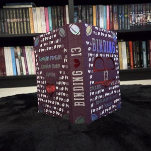 Puede incluir: Una caja decorativa con forma de libro con el título "BINDING 13" en letras blancas. La caja es de color morado oscuro con texto blanco y gráficos de corazón. La caja está sobre una superficie negra.