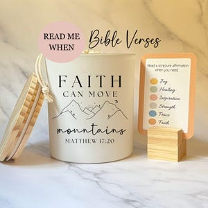May include: A white candle with the text "FAITH CAN MOVE mountains" and a mountain illustration. Includes a wooden lid and a card with scripture affirmations. The text "READ ME WHEN" is above the candle.
