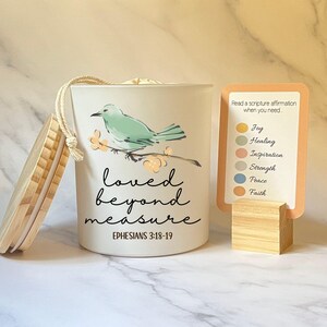 May include: A white candle with a wooden lid and a bird illustration. The candle has the words "loved beyond measure" and "Ephesians 3:18-19" printed on it. A card with scripture affirmations is displayed next to the candle.