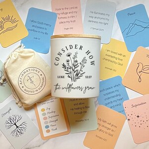 May include: A white candle with a wooden lid, featuring the text "CONSIDER HOW the wildflowers grow" and a floral design. Affirmation cards and a small bag with the text "AFFIRMATIONS ROOTED IN SCRIPTURE" surround the candle. The cards have inspirational quotes.