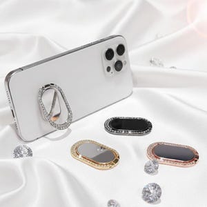 Peut inclure: Un smartphone blanc avec un support de bague argenté orné de strass. Trois accessoires de téléphone supplémentaires en or, noir et or rose, chacun avec une bordure en strass, sont présentés sur une surface en satin blanc. Cristaux décoratifs éparpillés.