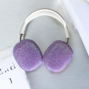 May include: A pair of wireless headphones with a white headband and ear cups covered in sparkling purple rhinestones. The headphones have silver-colored metal accents and are designed for over-the-ear use. The overall aesthetic is glamorous and stylish.