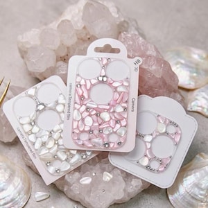 Könnte beinhalten: Drei dekorative Handy-Kameraschutzfolien. Eine ist rosa mit weißen Akzenten, die andere weiß mit silbernen Akzenten und die dritte rosa mit silbernen Akzenten. Jede hat ein einzigartiges Muster aus kleinen, unregelmäßigen Formen.