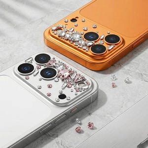 May include: Two bejeweled phone cases, one silver and one orange, are displayed. The silver case features pink and clear rhinestones around the camera lenses. The orange case has clear rhinestones. Loose rhinestones are scattered around the cases.
