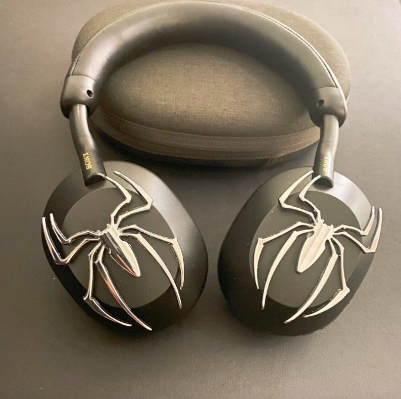 May include: Black wireless headphones with a silver spider design on each earcup. The headphones are on a gray surface.