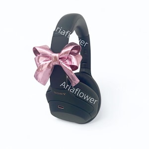 May include: Black over-ear headphones with a pink bow tied around the headband. The headphones have the word "SONY" printed on the side.