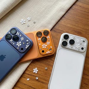 以下が含まれることがあります： 青、オレンジ、シルバーの装飾的な携帯電話ケース3つ。各ケースには、白い花とクリスタルのアクセントで飾られたカメラレンズプロテクターが付いています。携帯電話は、ベージュの布が付いた木製の表面に配置されています。
