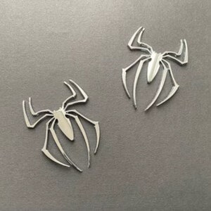 May include: Two silver-colored spider-shaped metal emblems. The emblems are detailed and have a shiny finish.