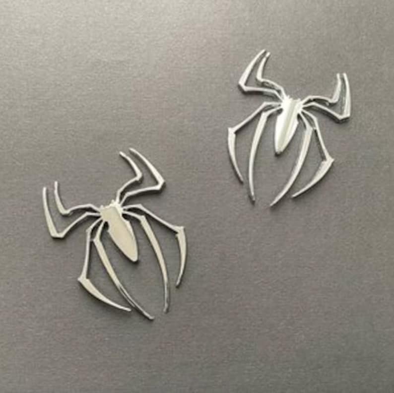 May include: Two silver-colored spider-shaped metal emblems. The emblems are detailed and have a shiny finish.