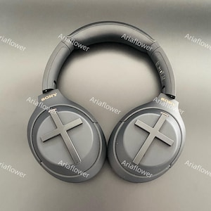 Puede incluir: Auriculares negros de diadema con diadema y almohadillas acolchadas. Los auriculares tienen un diseño de cruz plateada en cada auricular y la palabra "SONY" está impresa en el lateral de cada auricular.