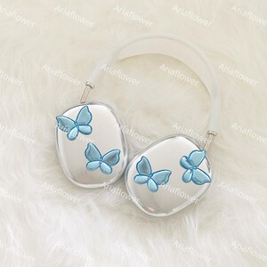 Peut inclure: Casque audio argent&eacute; avec un bandeau blanc et des papillons bleus clairs. Les &eacute;couteurs ont une surface r&eacute;fl&eacute;chissante, chacun avec deux ou trois d&eacute;corations de papillons. Le casque repose sur une surface blanche et duveteuse.