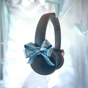 May include: Dark grey Sony headphones adorned with a metallic blue bow. The headphones are suspended from a pale green hook. The bow is positioned centrally, enhancing the headphones' appearance.