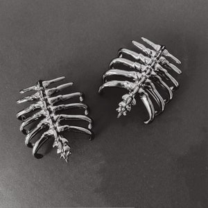 May include: Two silver skeleton ribcage hair clips. The clips are made of metal and have a detailed ribcage design.